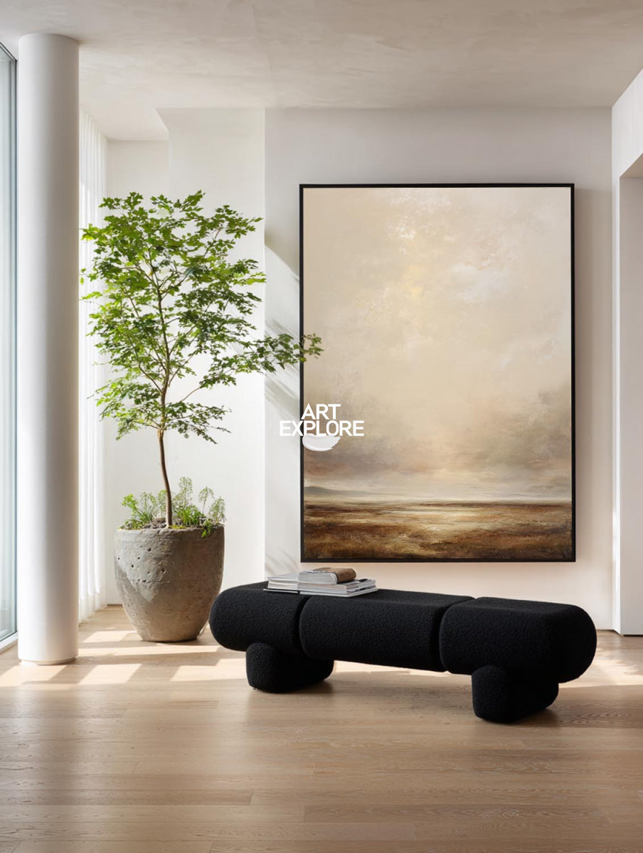 A large framed abstract landscape painting in beige, cream, and brown tones hangs on a white wall in a modern interior. Below the artwork is a black upholstered bench, and to the left is a tall potted tree in a concrete planter on a light wood floor.