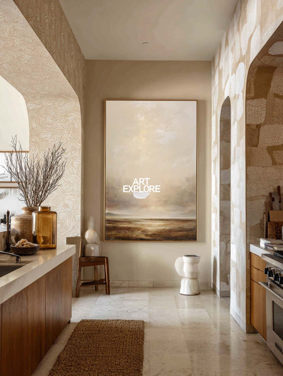 A large vertical landscape painting with soft beige, cream, and brown tones hangs on a wall in a modern kitchen with stone and wood elements.