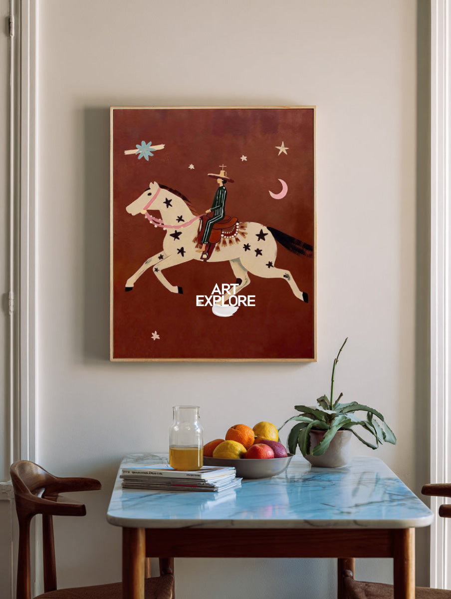 A framed western-style art print hangs on a beige wall above a dining table with fruit, books, and a plant. The artwork shows a rider on a horse against a rust-brown background with stars and a crescent moon.