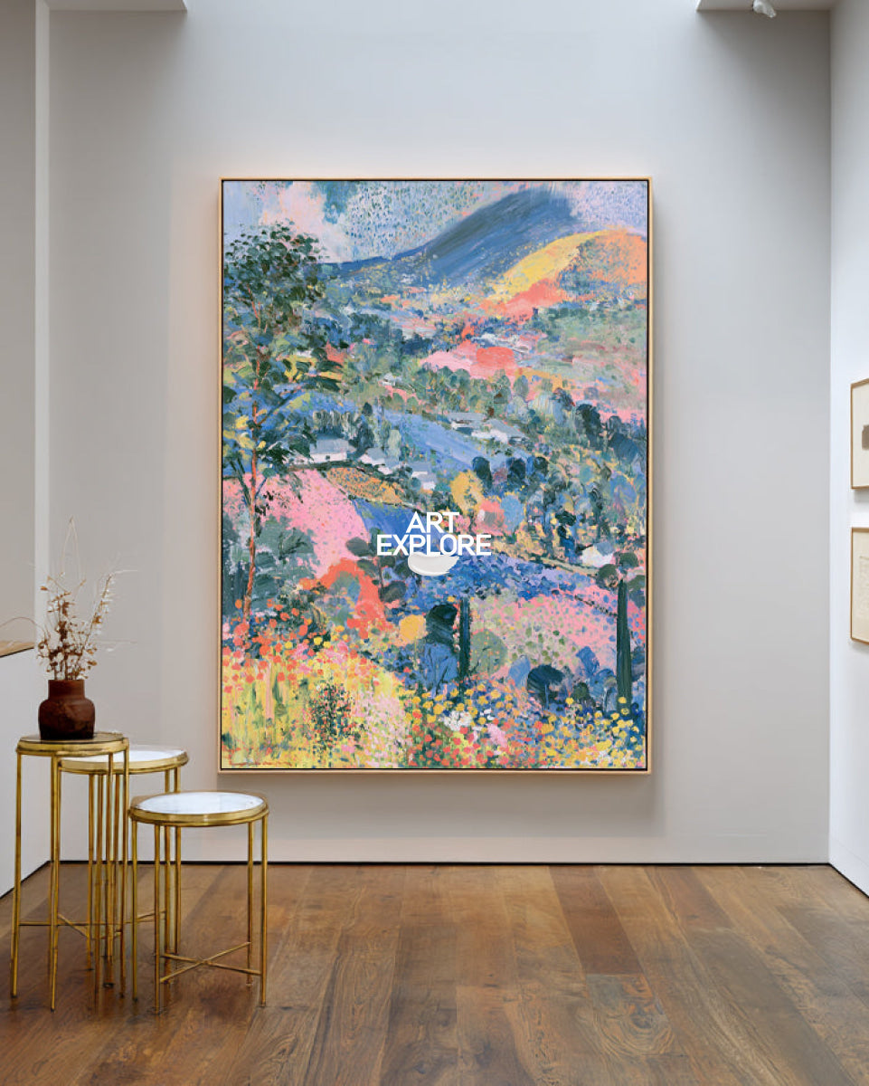 A colorful landscape painting in a gold frame, displayed on a light gray gallery wall above a wooden floor, with two gold side tables and a small vase in the foreground.