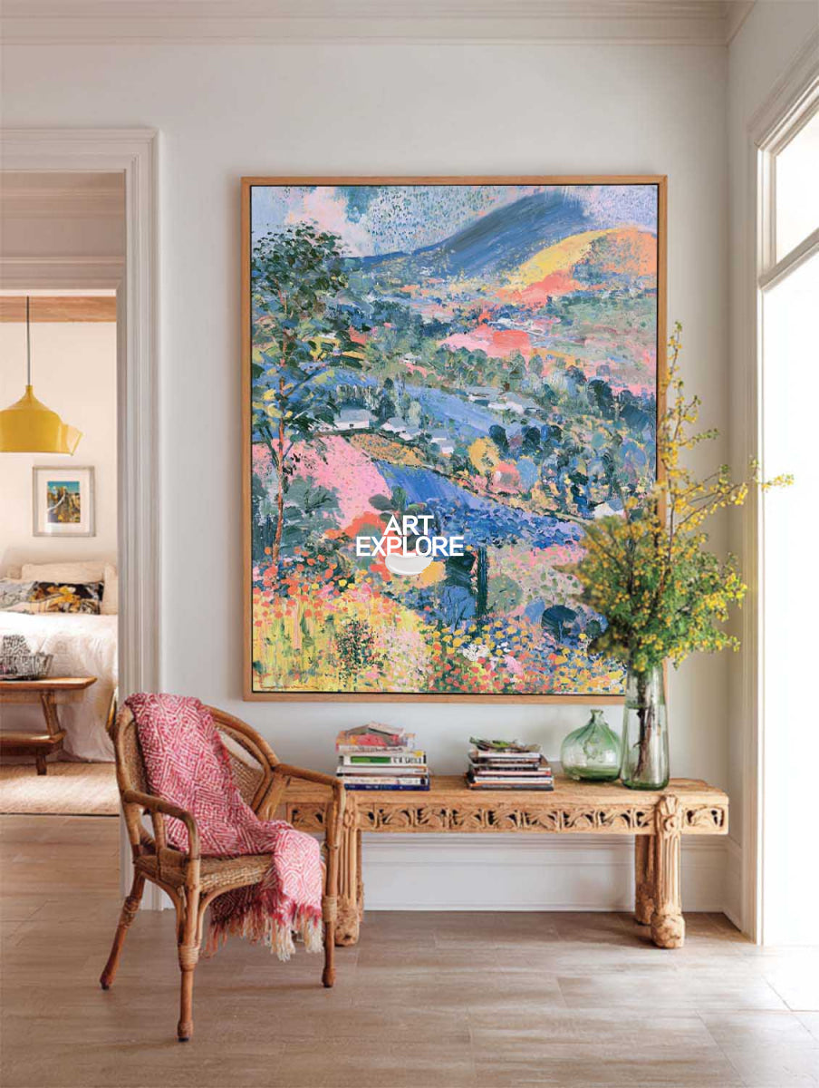 A large framed landscape painting with vibrant blue, green, pink, and yellow brushstrokes hangs on a light gray wall above a wooden bench in a styled room with a chair and vase.