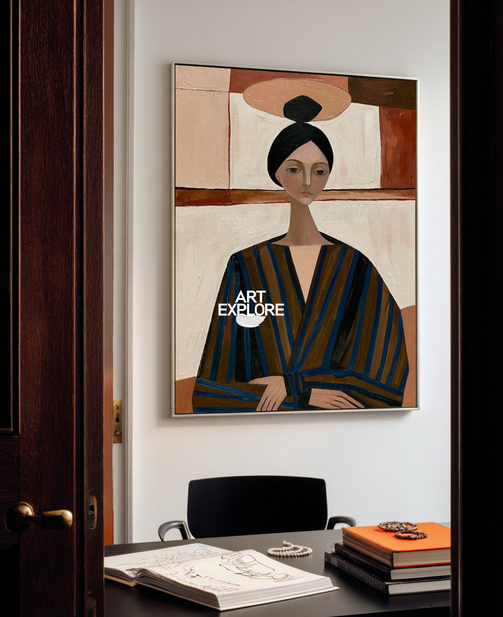 A framed art print featuring a stylized portrait of a woman in a striped robe, displayed on a white wall above a desk with books and an open sketchbook.
