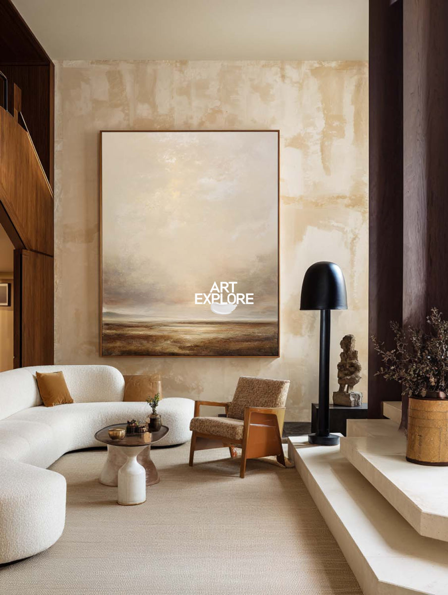 A large vertical landscape painting of a beach or sea under a cloudy sky, displayed on a textured beige wall in a modern, warm-toned living room with a curved white sofa, wooden chair, and black floor lamp.
